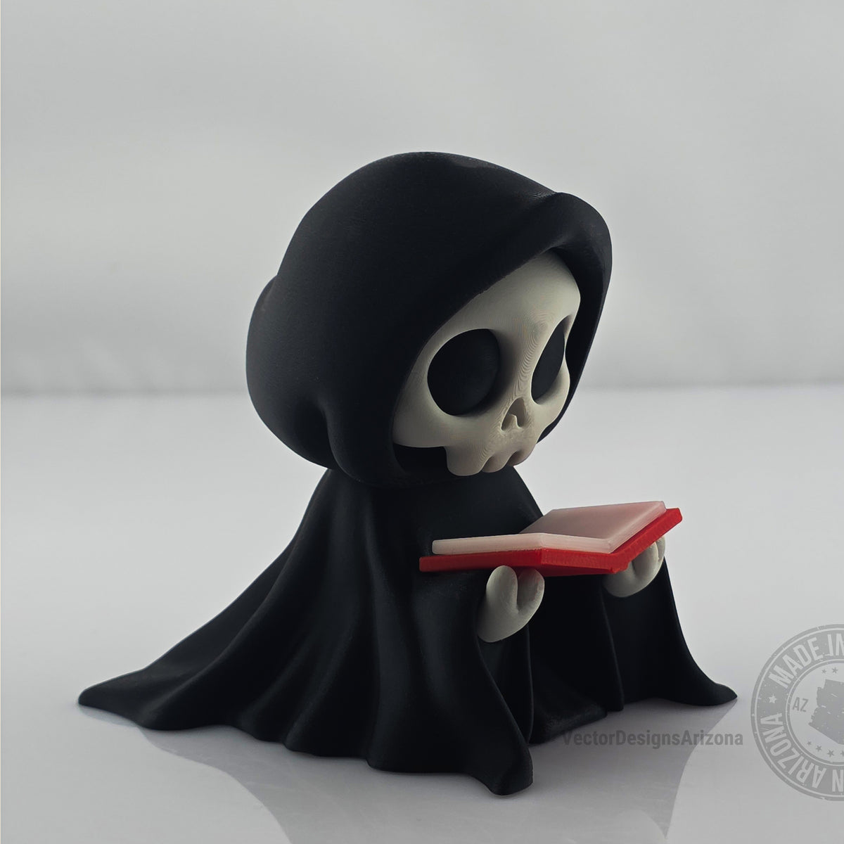 Cute hooded grim reaper figurine reading a small red-edged book on white background.