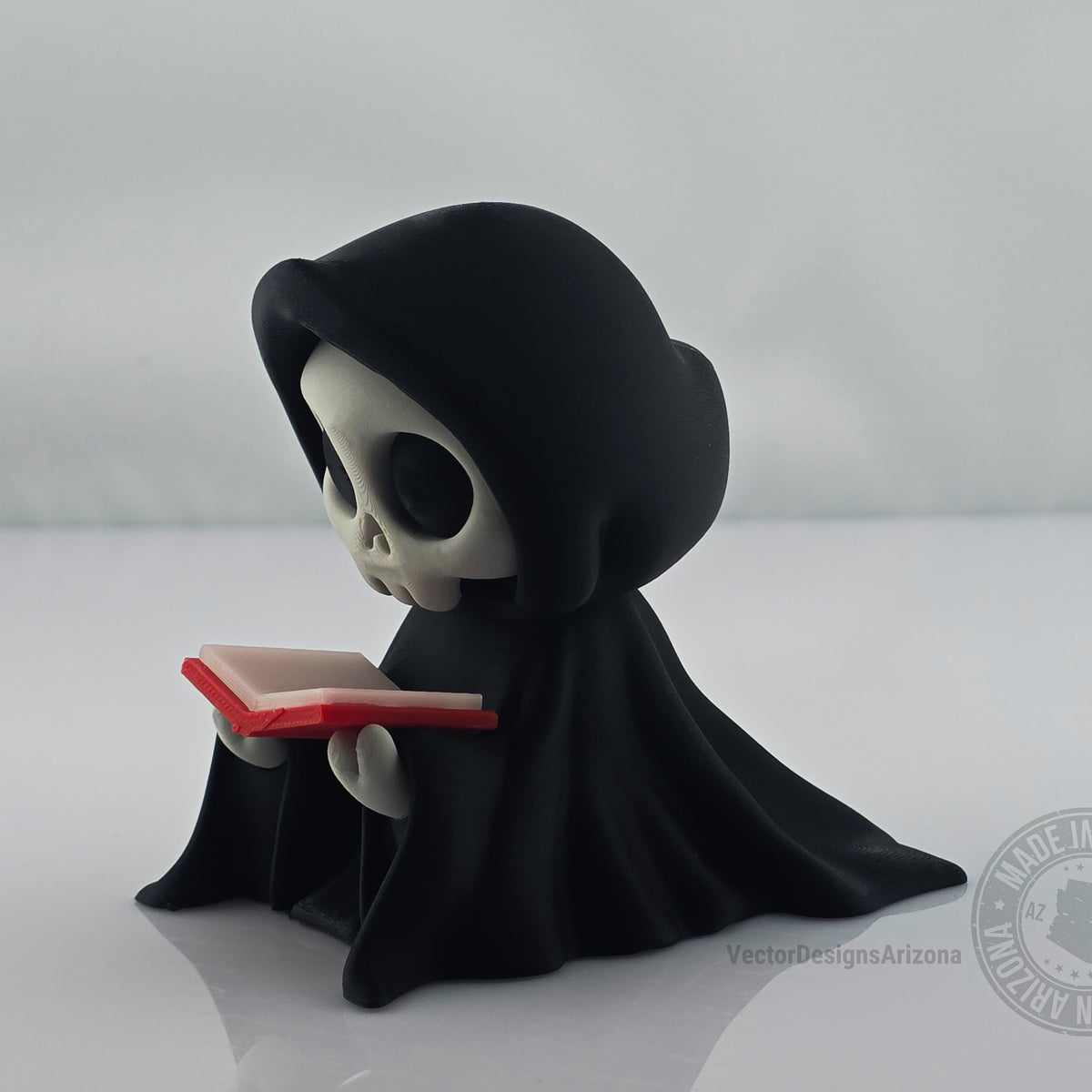 Side view of cute grim reaper figurine holding red-edged book on white background.