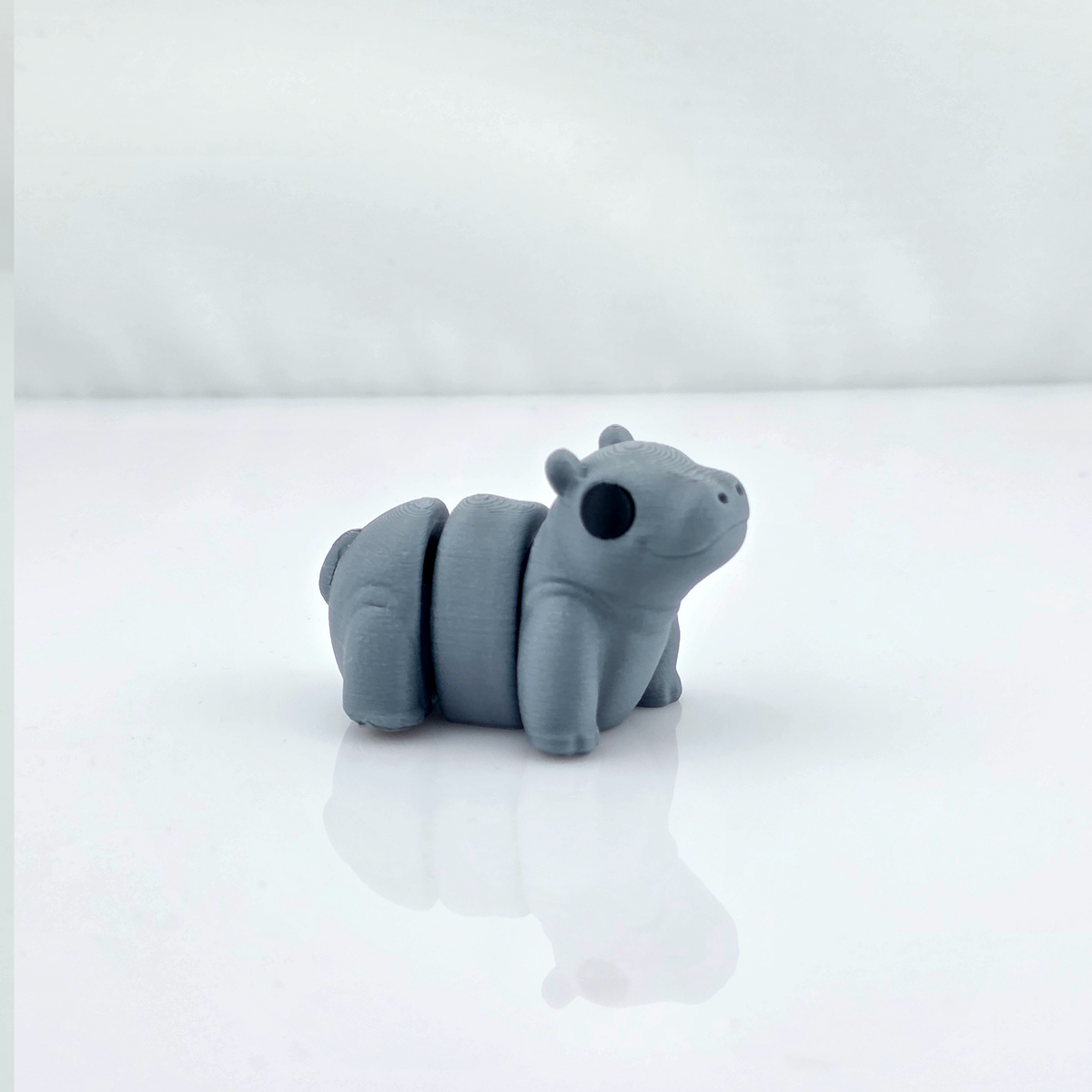 Gray mini flexy hippo toy standing on glossy white background with reflection.