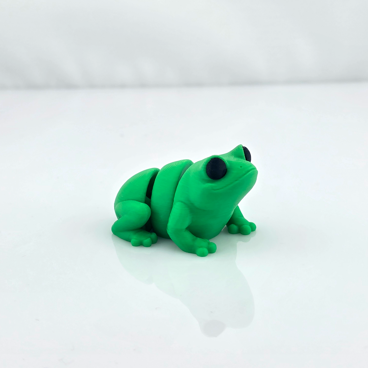 Green mini flexy frog toy crouching on glossy white background, three-quarter view.
