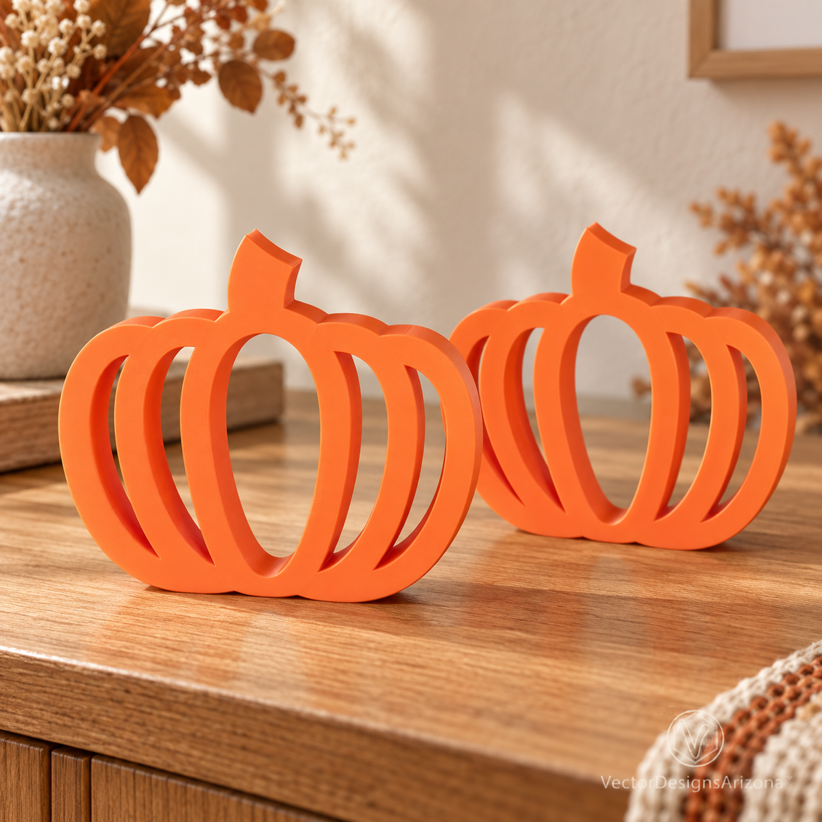 Two Mandarin orange minimalist 3D-printed pumpkins with open-frame designs, styled on a wood console with warm fall decor.