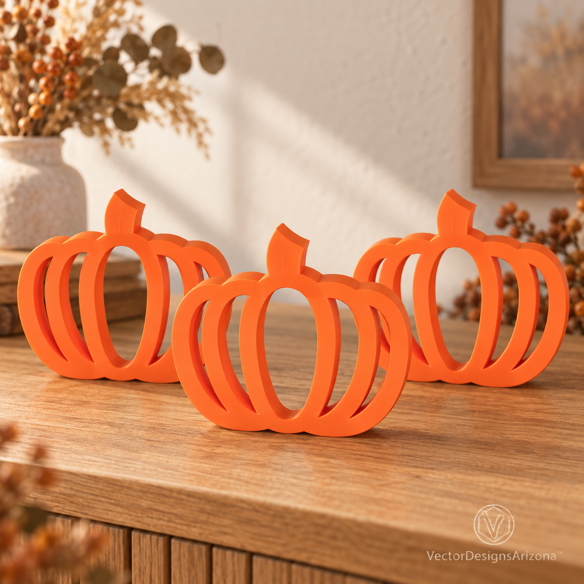 Three Mandarin orange minimalist 3D-printed pumpkins with open-frame designs, styled on a wood console with warm fall decor.
