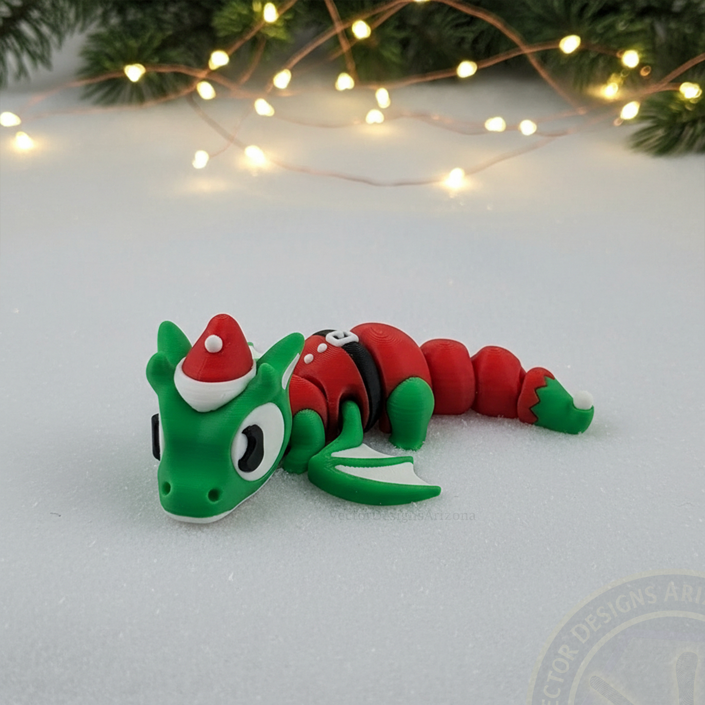 VectorDesignsArizona 3D printed flexi Elf Dragon Christmas figurine resting on snow with warm holiday lights in the background.