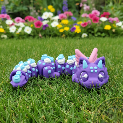 Purple Blueberry Dragon 3D printed flexi fidget toy posed on green grass with colorful flowers blurred in the background.