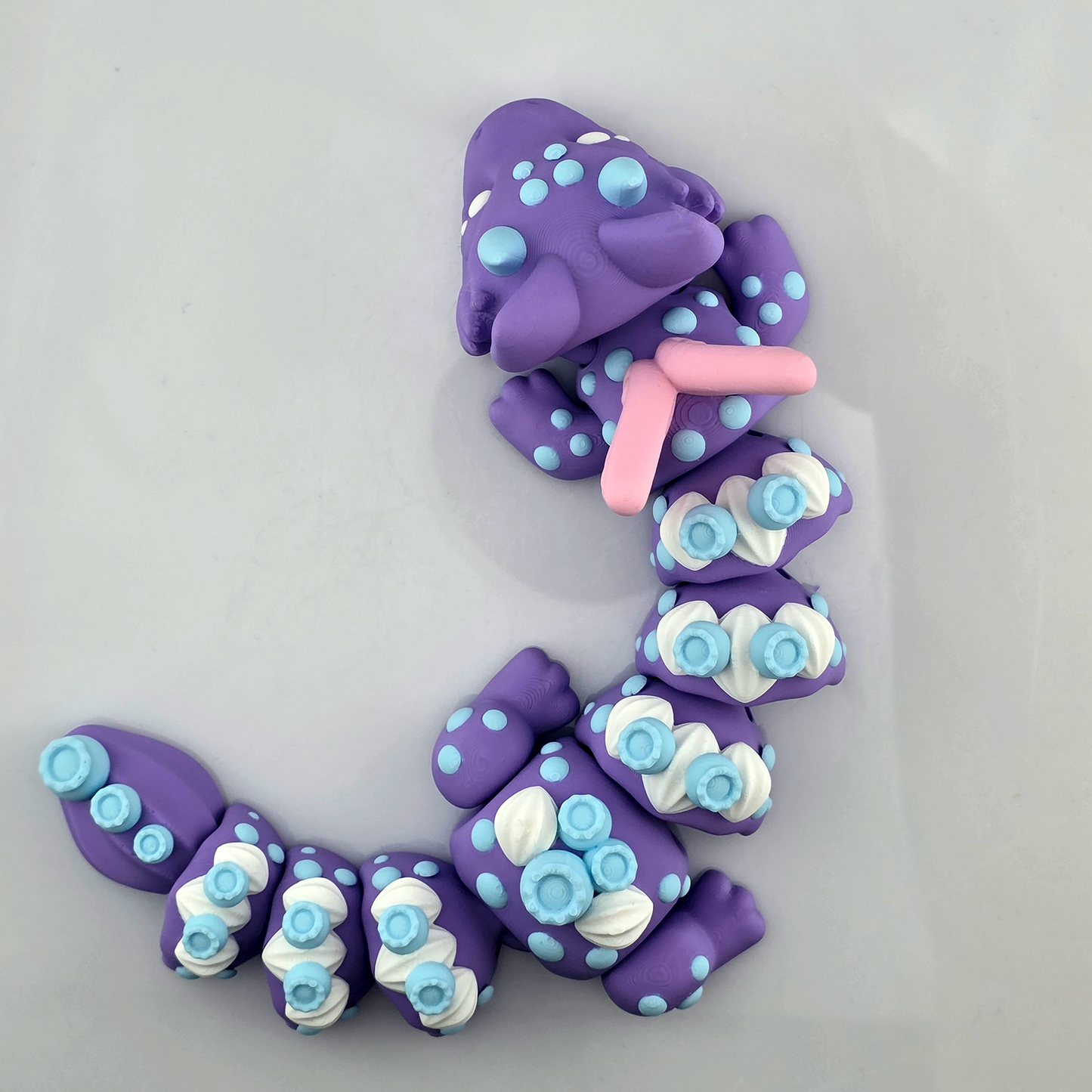 Top view of a purple Blueberry Dragon 3D printed flexi fidget toy with blue spots and pink wings on a white background.