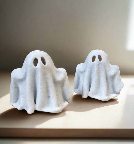 Two minimalist white ghost figurines on neutral surface in soft natural light.