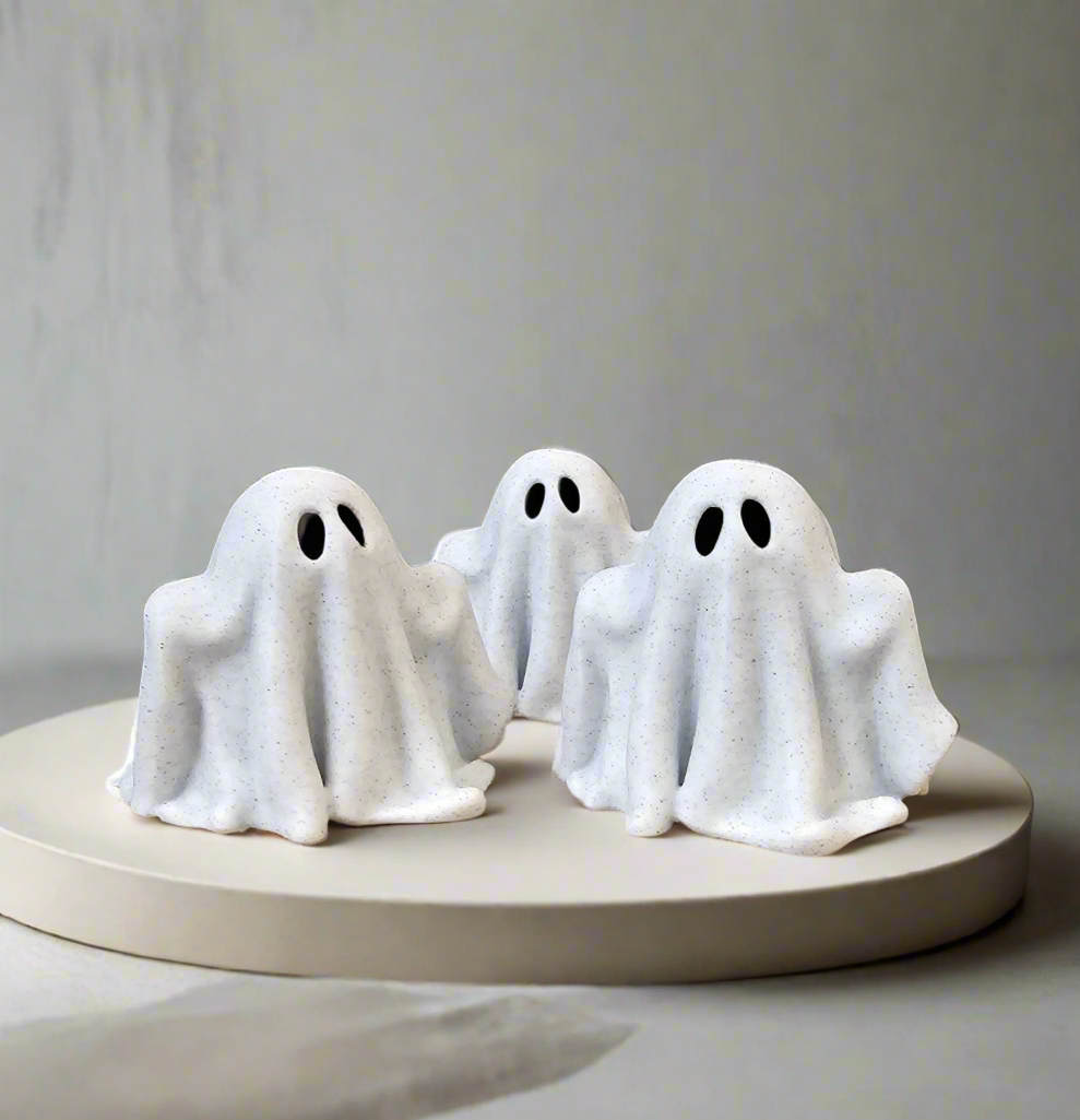 Three minimalist white ghost figurines arranged on a round neutral pedestal.