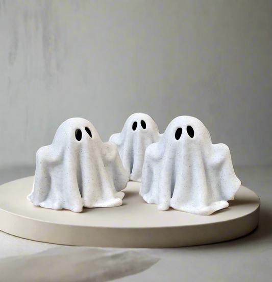 Three minimalist white ghost figurines arranged on a round neutral pedestal.