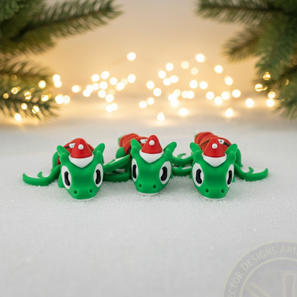 Three VectorDesignsArizona 3D printed flexi Elf Dragon Christmas figurines lined up on snow with pine branches and warm lights behind them.