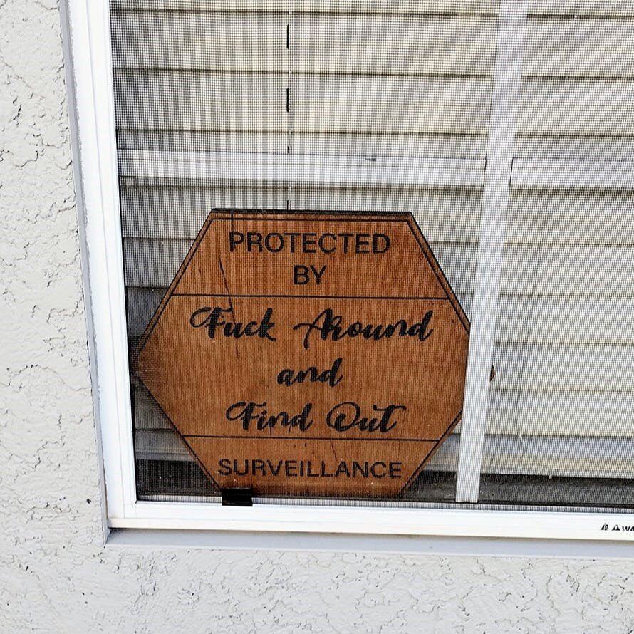 Wood FAFO surveillance sign displayed inside house window behind screen and blinds.
