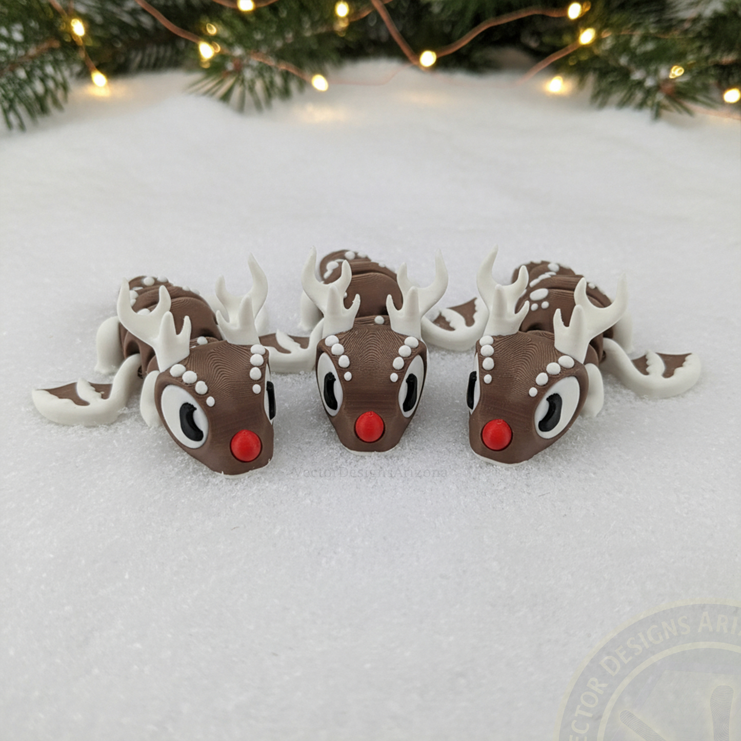 Three VectorDesignsArizona 3D printed flexi Reindeer figurines lined up on snow in front of pine branches and holiday lights.