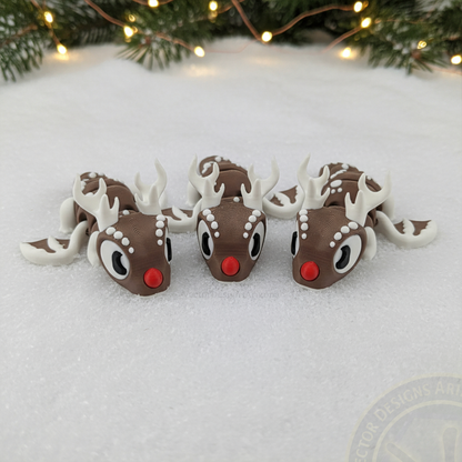 Three VectorDesignsArizona 3D printed flexi Reindeer figurines lined up on snow in front of pine branches and holiday lights.