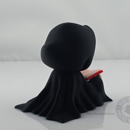 Back side view of cute grim reaper figurine reading a red-edged book.