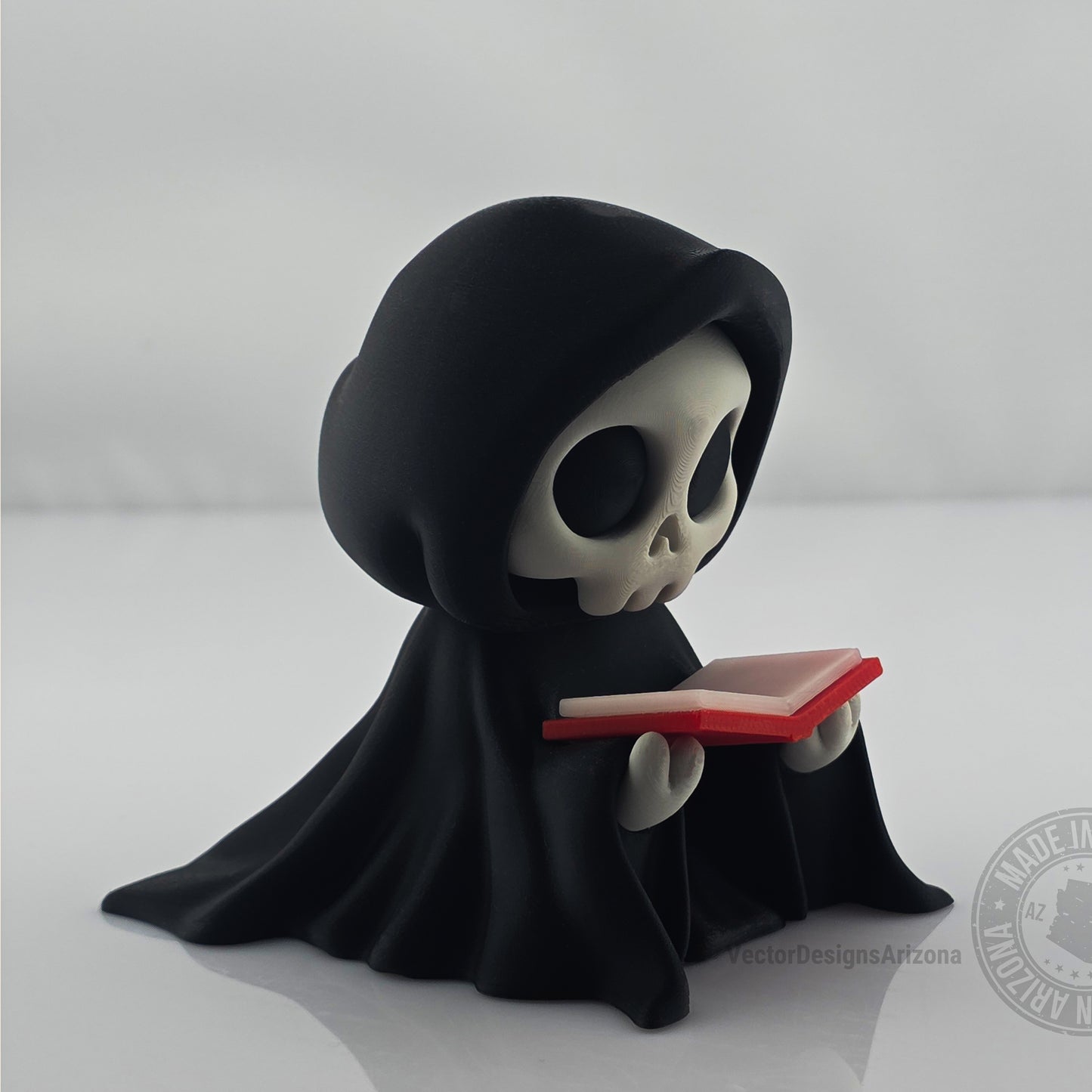 Cute hooded grim reaper figurine reading a small red-edged book on white background.