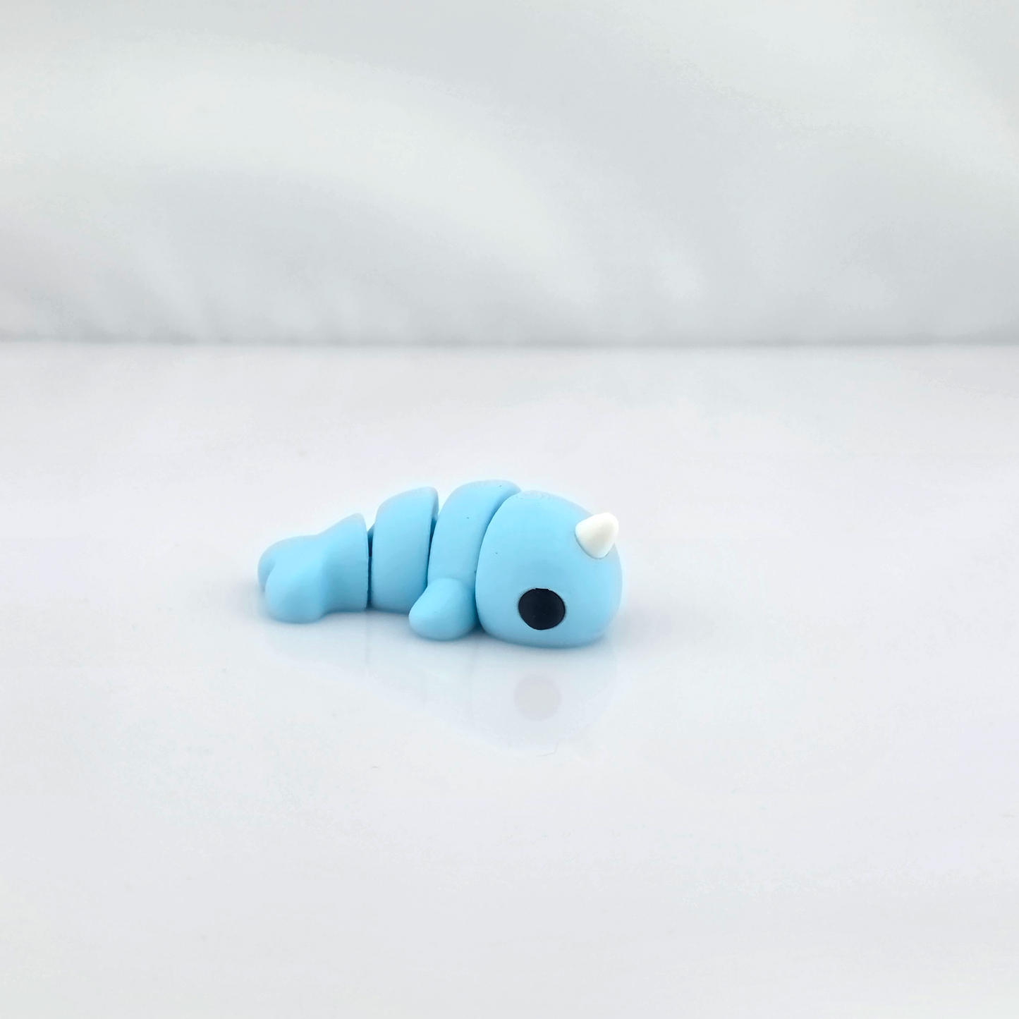 Light blue mini flexy narwhal toy lying on glossy white background.