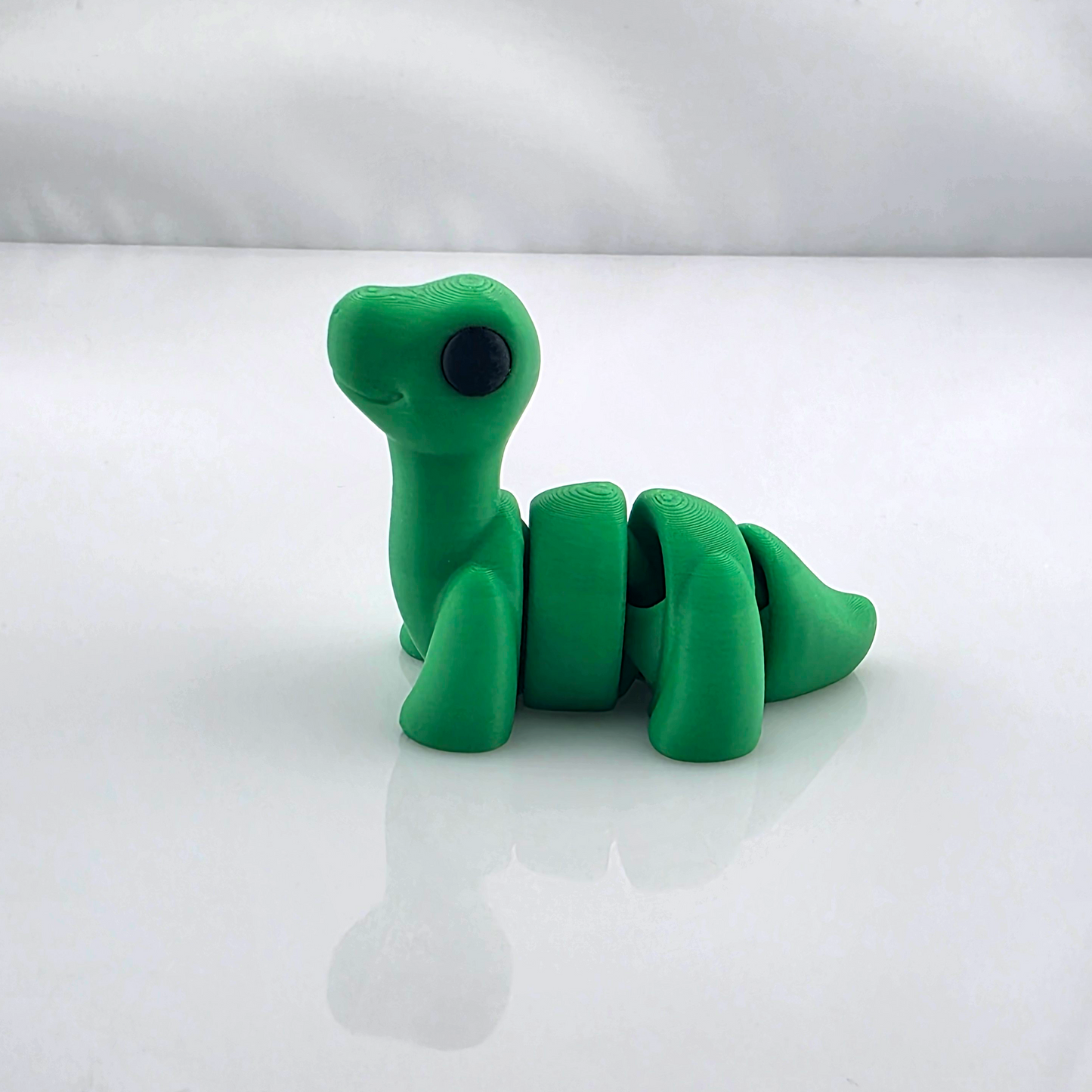 Green mini flexy dinosaur toy sitting upright on glossy white background.
