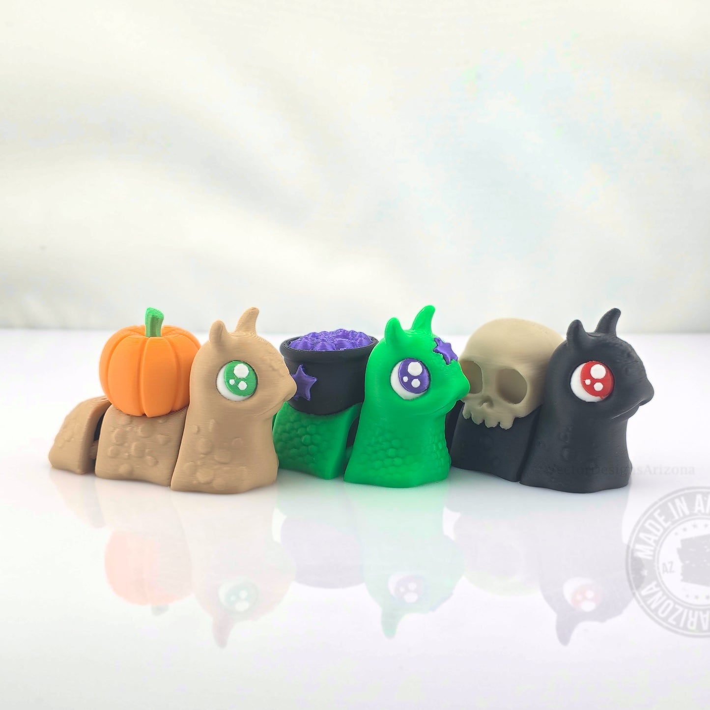Pumpkin, cauldron, and skull spooky snail figurines lined up on white reflective background.