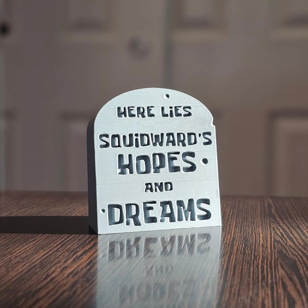 Small gray tombstone desk sign on wooden surface with humorous engraved text.