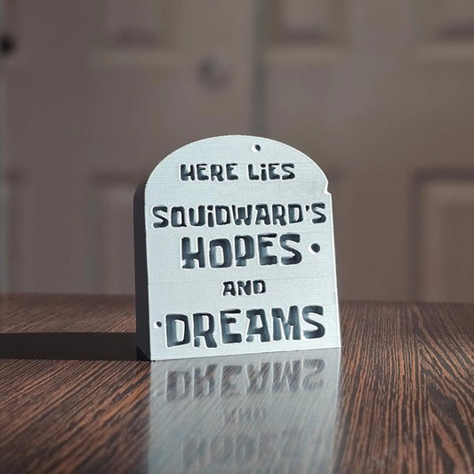 Small gray tombstone desk sign on wooden surface with humorous engraved text.